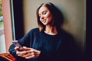 Cheerful young woman using free high speed internet connection for chatting online on smartphone device.Positive hipster girl laughing and installing application on mobile phone resting in coworking