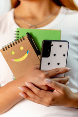  A woman is holding a cell phone and a notebook with a smiley face on it. She is wearing a white...