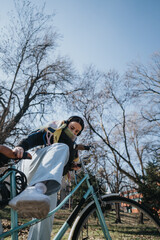 Young female cyclist with headphones rides leisurely in a city park, enjoying a sunny day amidst...
