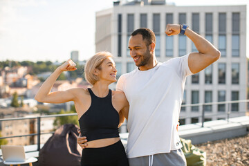 Young couple in sportswear poses against background of urban buildings on roof. Smiling athletic man and woman showing biceps while exercising healthy lifestyle.