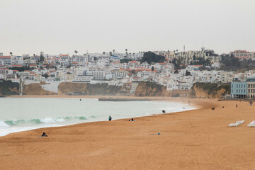 People Walking Along the Beach as Waves Crash in on During a Hazy Sunset in Albufeira Portugal