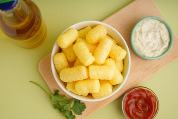 Bowl with tasty salty corn sticks, bottle of beverage and sauces on green background