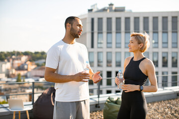 Portrait of young sportsman and fit woman in sportswear resting after running outdoors holding...