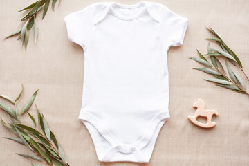 Mockup of white baby bodysuit on linen with greenery and toy. Blank baby clothes template in farmhouse style, flat lay.
