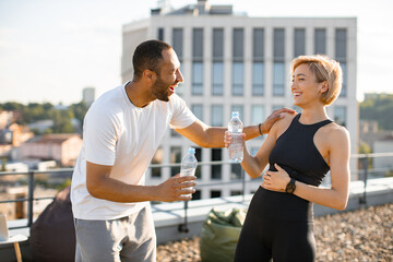 Athletic man in white t-shirt and woman in black top drinking water after jogging. Young couple having fun laughing after morning workout on the roof of the house.