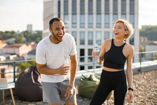 Athletic man in white t-shirt and woman in black top drinking water after jogging. Young couple having fun laughing after morning workout on the roof of the house.