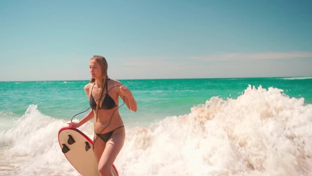 Woman holding surfboard rope while standing in sea waves on beach. Beautiful wet woman in black bikini is enjoying surfing on her summer vacation. 