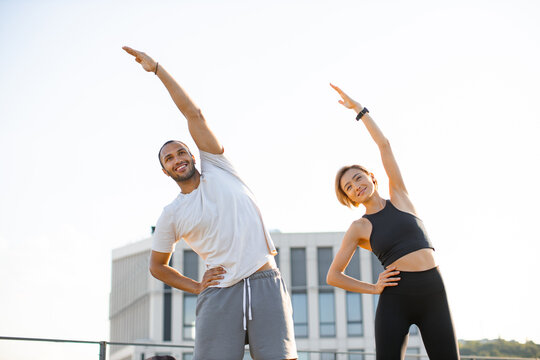 Happy sports couple making health gymnastics in morning on roof terrace of building. Positive man in white t-shirt and woman in black top doing sports stretching their arms up. - Powered by Adobe