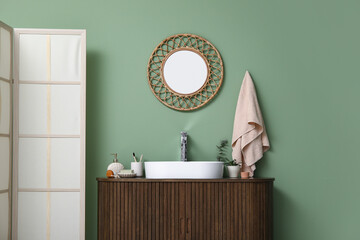 Modern sink with bath supplies on countertop and stylish mirror near green wall in bathroom