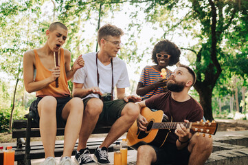An interracial group of students sitting in a park and playing guitar.