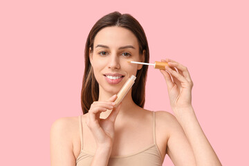 Beautiful young woman applying concealer on pink background