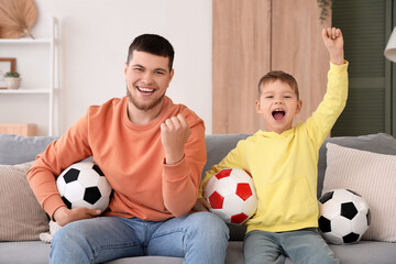 Young man and his little son cheering for football team with soccer balls in living room