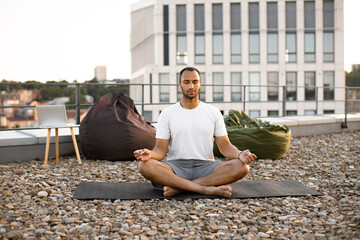 Young athletic man doing yoga at sunset on urban background. Handsome male meditating in lotus...