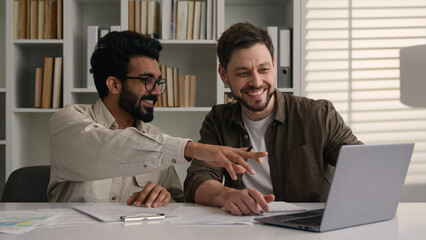 Two happy smiling multiethnic colleagues businessmen business partners at office company discuss project computer work Caucasian Indian men male coworkers look at laptop corporate teamwork discussion