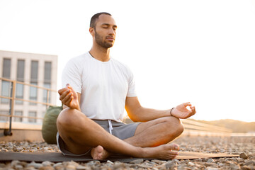 Handsome male meditating in lotus position while sitting on mat on roof of modern house. Young...
