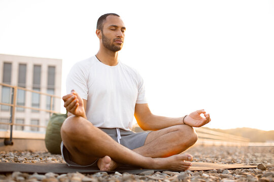 Handsome male meditating in lotus position while sitting on mat on roof of modern house. Young athletic man doing yoga at sunset on urban background.