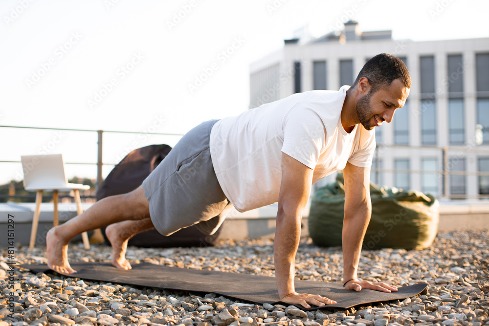 Sticker Young smiling sports man doing morning exercises on roof of modern house. Strong male in plank position on mat during sunrise against urban background.