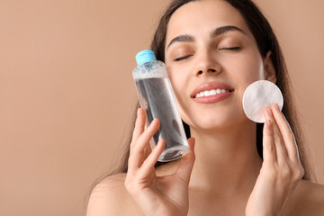 Beautiful young happy woman with cotton pad and bottle of micellar water on brown background
