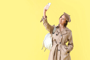 Happy female student with backpack taking selfie by mobile phone on yellow background