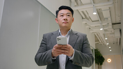 Middle-aged Asian Korean man thoughtful chinese businessman CEO employer in office hallway use phone mobile app browsing internet typing chatting message online in smartphone think choose solve ponder