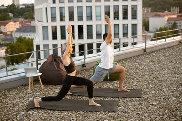 Concept of meditation and inner development of soul. Sporty family practicing warrior yoga pose with raised arms standing on mats on roof of modern house in summer morning.