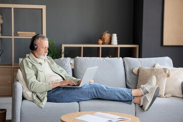 Mature man in headphones using laptop on sofa at home