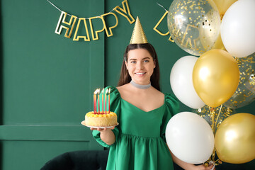 Young woman in party hat with Birthday cake and balloons near green wall