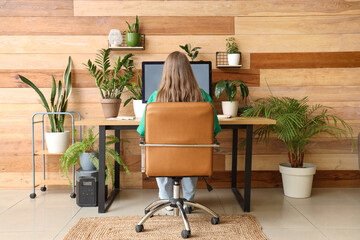 Young woman working with computer at table and plants near wooden wall in office, back view