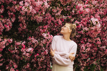 Young stylish woman in glasses surrounded by pink spring flowers hugging herself.