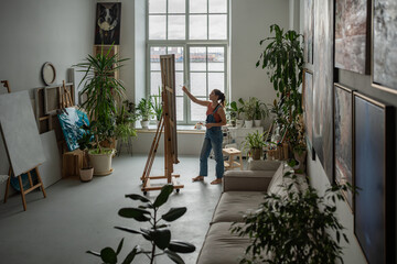 Creative female artist paints with oil on canvas, standing near easel with brush. Focused woman...