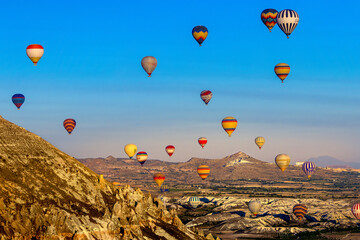 Naklejka premium Colorful sunrise with multiple hot air balloons over a rugged landscape, perfect for travel, adventure, and lifestyle themes.