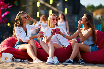 Group of joyful females on red bean bags clinking glasses at a beach music festival. They enjoy cold beverages, laugh in sunlight with festival stage in background. Casual summer gathering by sea.