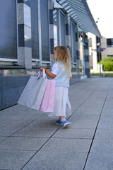 happy child of 3-4 years old go out of store, girl in light fashionable clothes, craft paper bags...