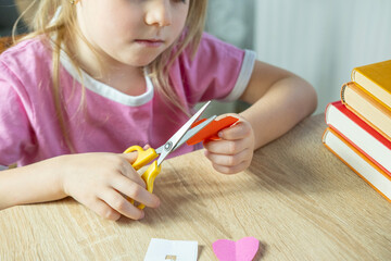 young girl learning to cut paper with scissors, making shapes like flowers for DIY art project,...
