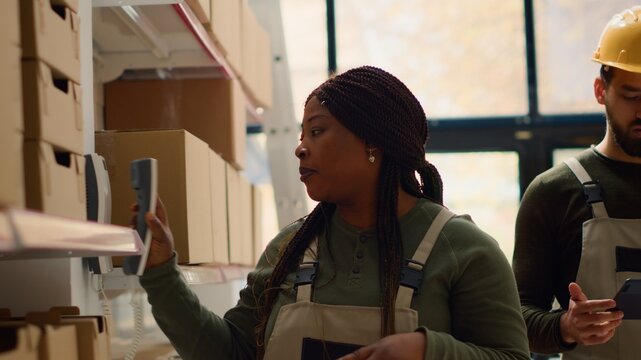 Logistics coordinator and warehouse picker preparing orders for delivery, receiving phone call in retail storehouse while scanning labels on cardboard packages to be shipped
