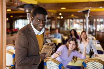 Cunning african american man trying to thieve handbag of young woman sitting at table in restaurant...