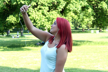 Seductive young redhead woman taking a selfie photo in public park while listening to music 