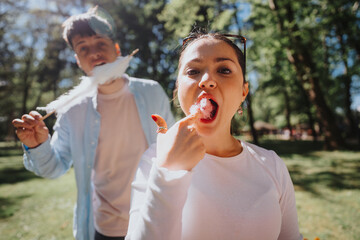 Fototapeta premium Two young friends savor cotton candy in a sunny park, sharing cheerful moments together with joyful expressions.