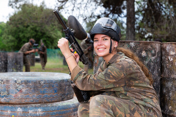 Fototapeta premium Young smiling woman paintball player in camouflage posing with gun outdoors
