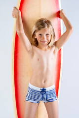 Cute young surfer boy standing in front of his surfboard smiling adorably