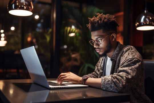 A black student using laptop and taking notes during an online class at a cozy coffee shop table during nighttime
