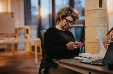 A mature businesswoman is concentrated on her phone during a break in a collaborative office space with colleagues around.