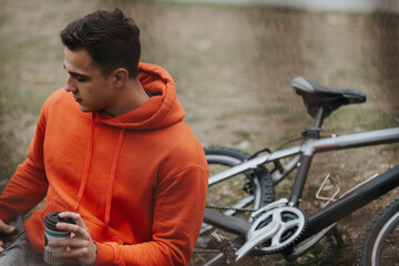 A casual young man sipping coffee next to his bicycle, relaxing in the park under a cloudy sky.
