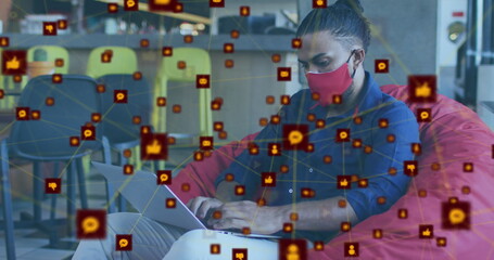 Image of network of digital icons over biracial man wearing face mask using laptop at office
