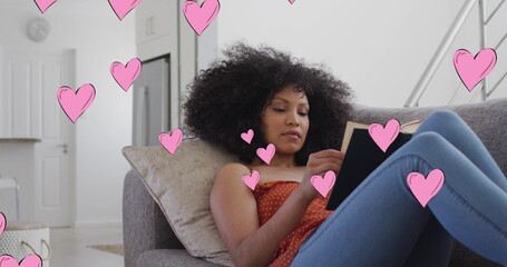 Image of social media heart icons over biracial woman reading book on sofa - Powered by Adobe