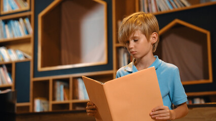 Attractive caucasian clever boy reading a book at library with stack of books placed. Smart child...
