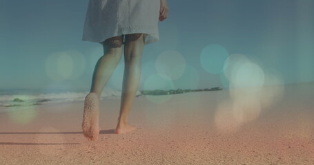 Image of lens flare over low section of biracial young woman walking barefoot on sandy beach - Powered by Adobe