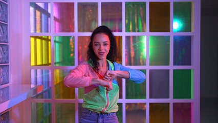 Happy hispanic girl in lively mood moving along music in neon light while wearing colorful cloth at...