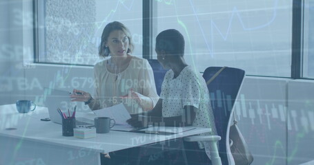 Image of graphs and trading board over diverse female coworkers discussing reports on laptop