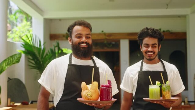 Two bipoc smiling waiters in aprons carry drinks to multiracial women in cafe on Bali. Friendly waiters put beverages on table, talk with female customers in a restaurant. Cafe workers serve clients.